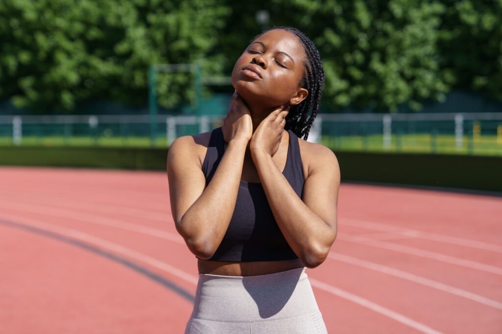 Sportive African American woman puts hands on stiff neck leaning head to sides on blurred background