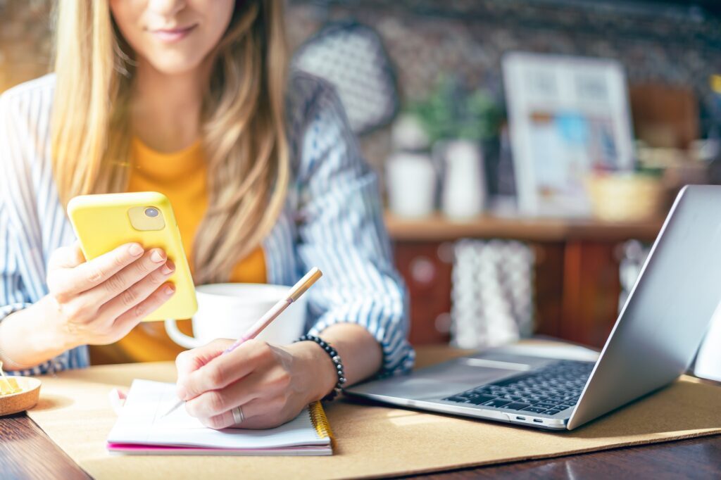 Woman with phone writing in notebook. Working home online, using technology laptop and cellphone.